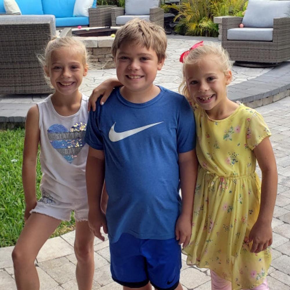 Kaleb, 10, stands between his 8-year-old twin sisters. All three smile at the camera.
