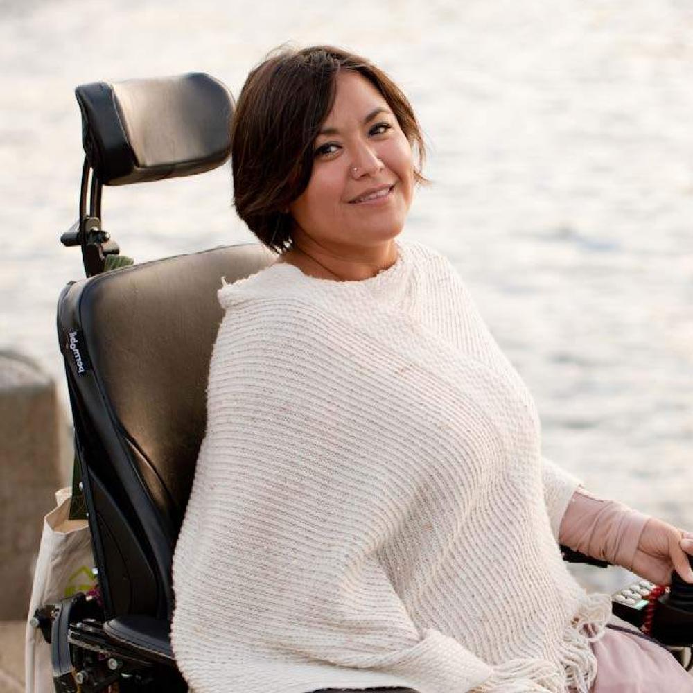 A 40-something woman in a white wrap sits in a power wheelchair in front of water. She looks up at the camera with a slight smile.