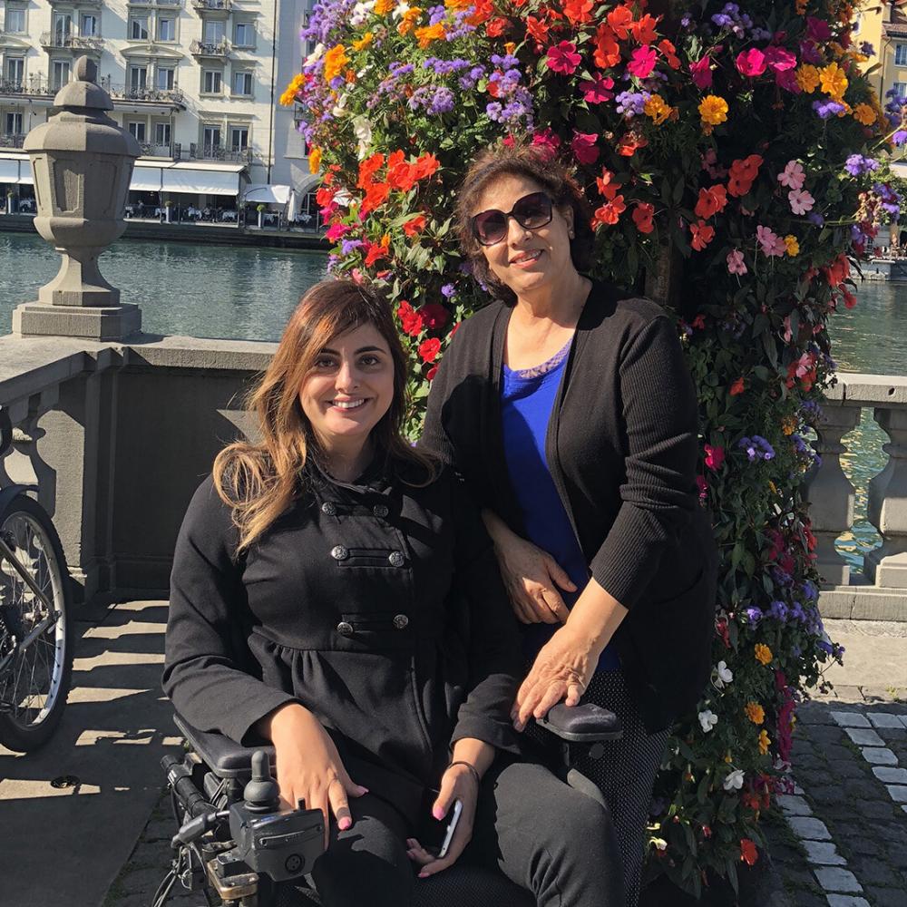 An older mother stands next to her 30-something daughter who is in a wheelchair. They are standing on a walkway in front of a river, smiling at the camera.